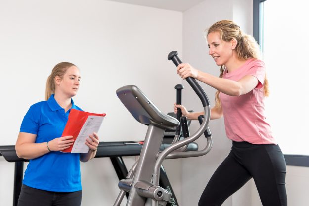 Bewegen Een vrouw is aan het bewegen op een crosstrainer. De fysiotherapeut staat naast haar en heeft een map in haar handen.