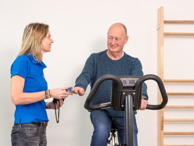 Man in zijn pensioen traint op een hometrainer onder begeleiding van een fysiotherapeut.