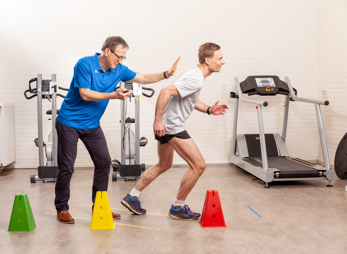 Patiënt in een wit shirt werkt aan sterke benen met behulp van de fysiotherapeut in een blauw shirt in de oefenzaal