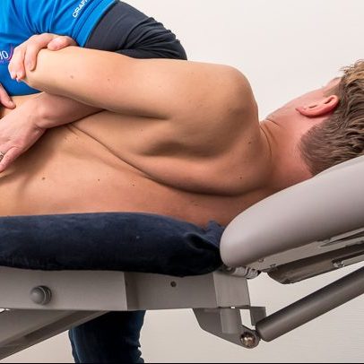 Patiënt zonder shirt wordt geholpen aan spit door de fysiotherapeut in een blauw shirt op de behandelbank