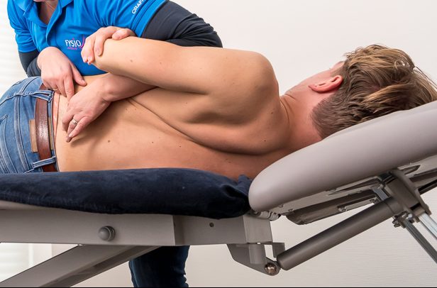 spit – Fysiotherapie Zesgehuchten Patiënt zonder shirt wordt geholpen aan spit door de fysiotherapeut in een blauw shirt op de behandelbank