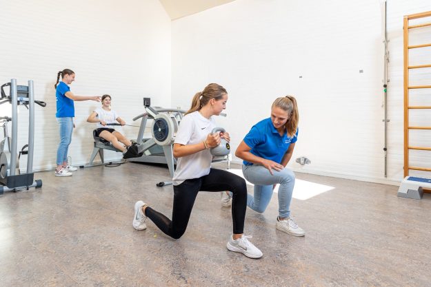 Sportieve vrouw die een kettlebell vasthoudt voorkom shin splints in een wit shirt en zwarte sportbroek werkt met behulp van de fysiotherapeut in een blauw shirt
