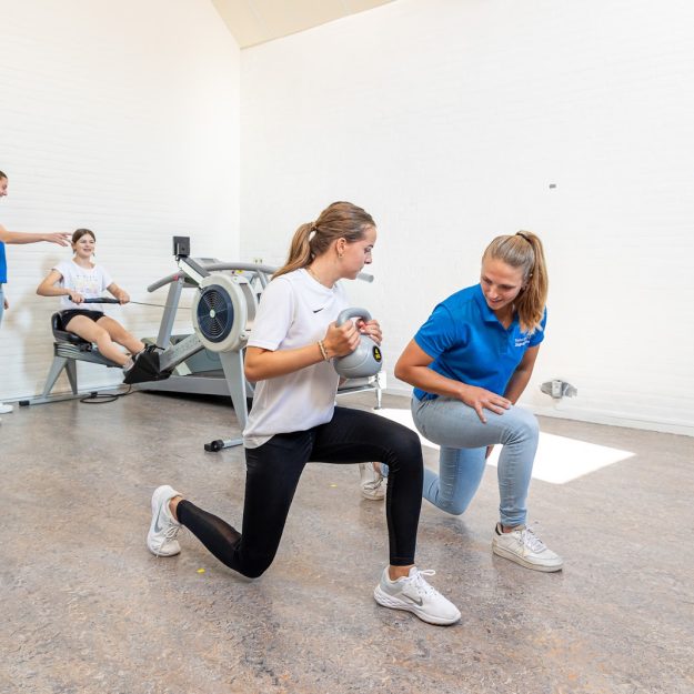 Sportieve vrouw die een kettlebell vasthoudt voorkom shin splints in een wit shirt en zwarte sportbroek werkt met behulp van de fysiotherapeut in een blauw shirt