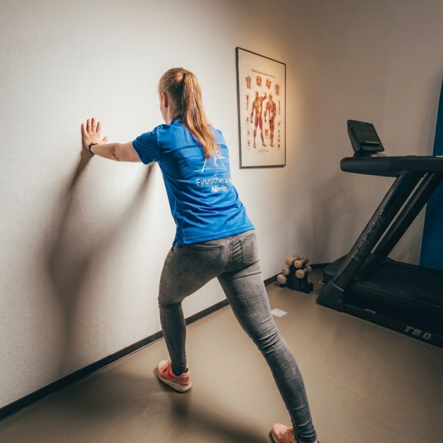 Fysiotherapeut staat tegen een muur en laat zien hoe je dagelijks moet stretchen.